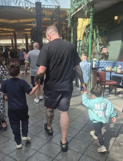 Man with arm tattoos holds hands with two young children, walking in a mall.