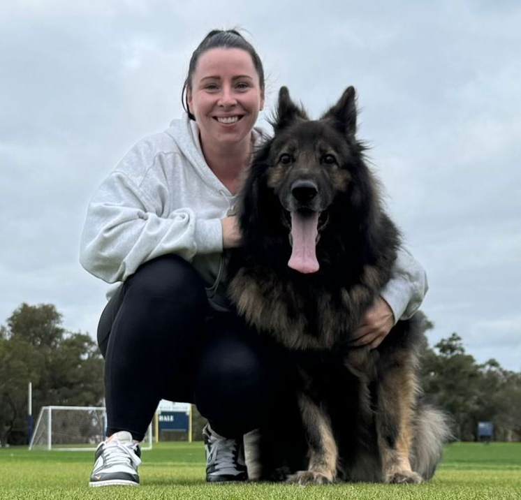 Woman in a gray hoodie and black leggings kneels next to a large, long-haired dog on a grassy field.