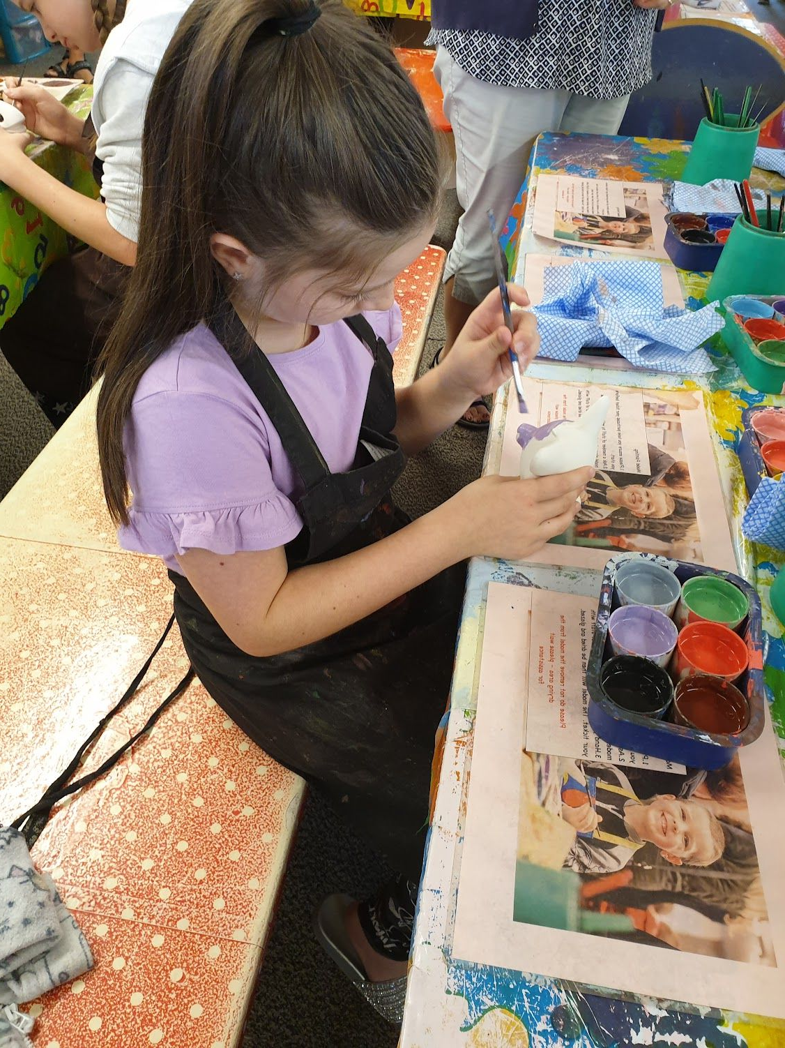 Girl painting at a table, wearing an apron, smiling. Paint colors and supplies nearby.