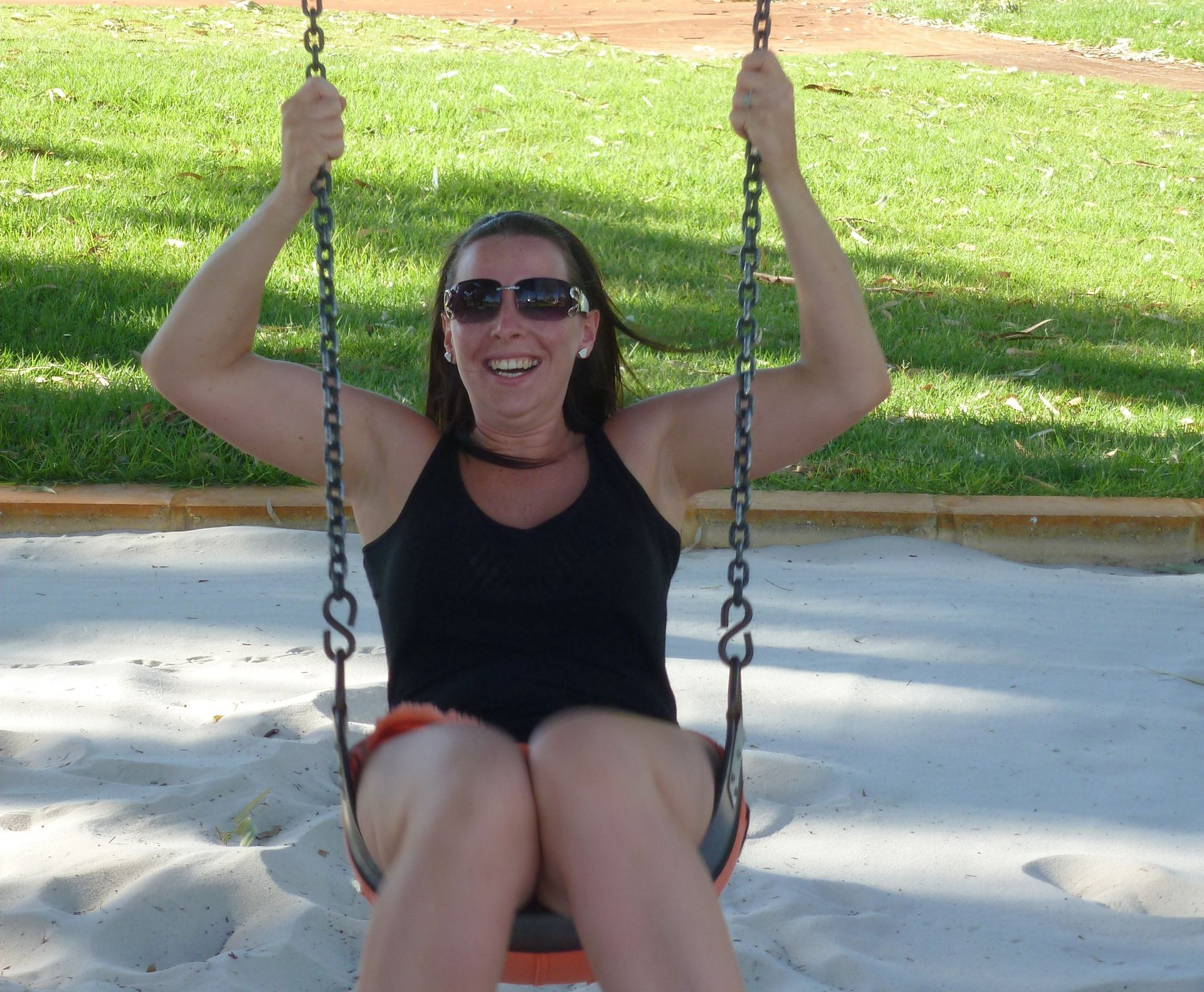 Woman on swing, laughing. Sunglasses, black top, shorts. Playground setting with sand, trees.