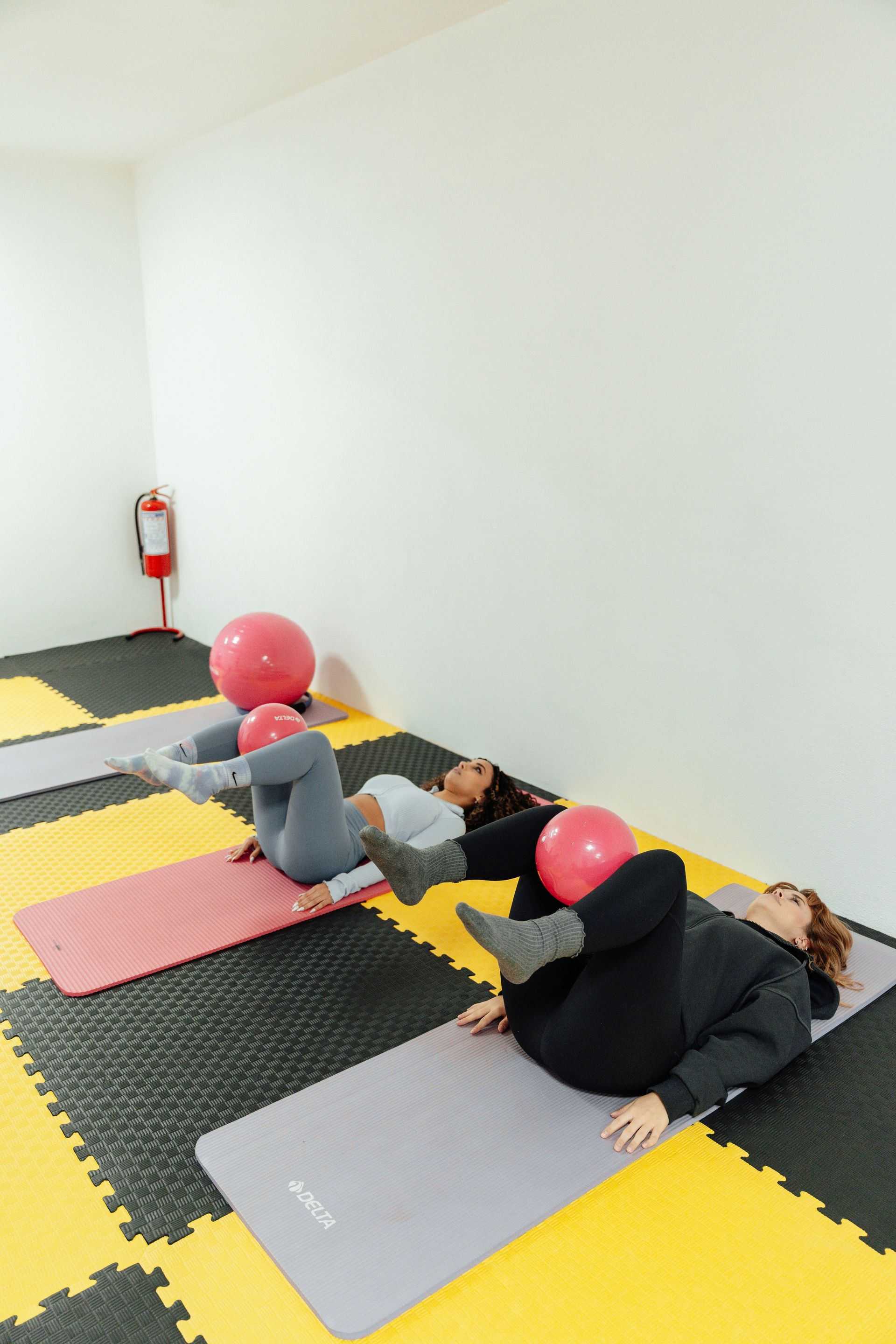 Three women on mats, exercising with pink balls in a room with yellow and black floor tiles.