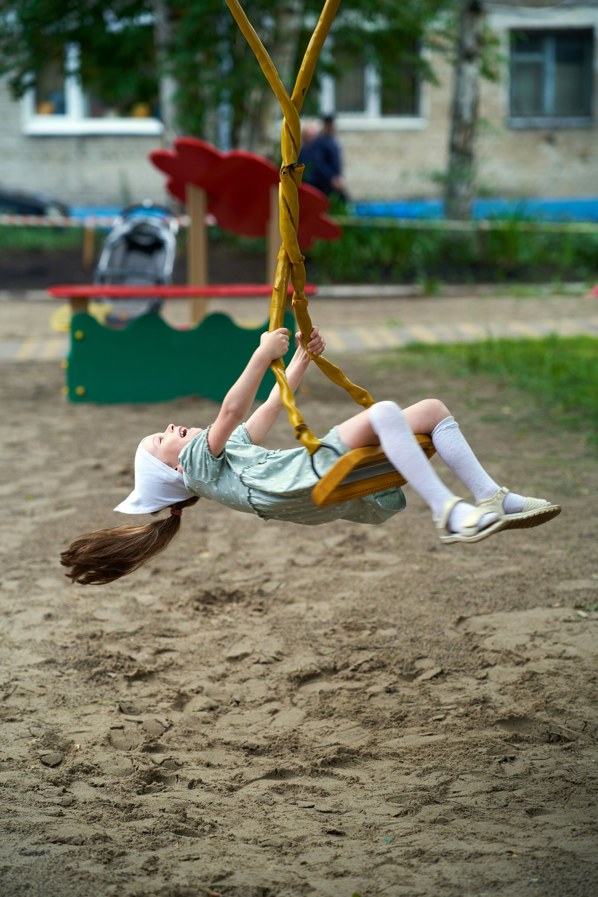 Girl on a swing at a playground, laughing, brown hair down, white hat, green dress, and socks.