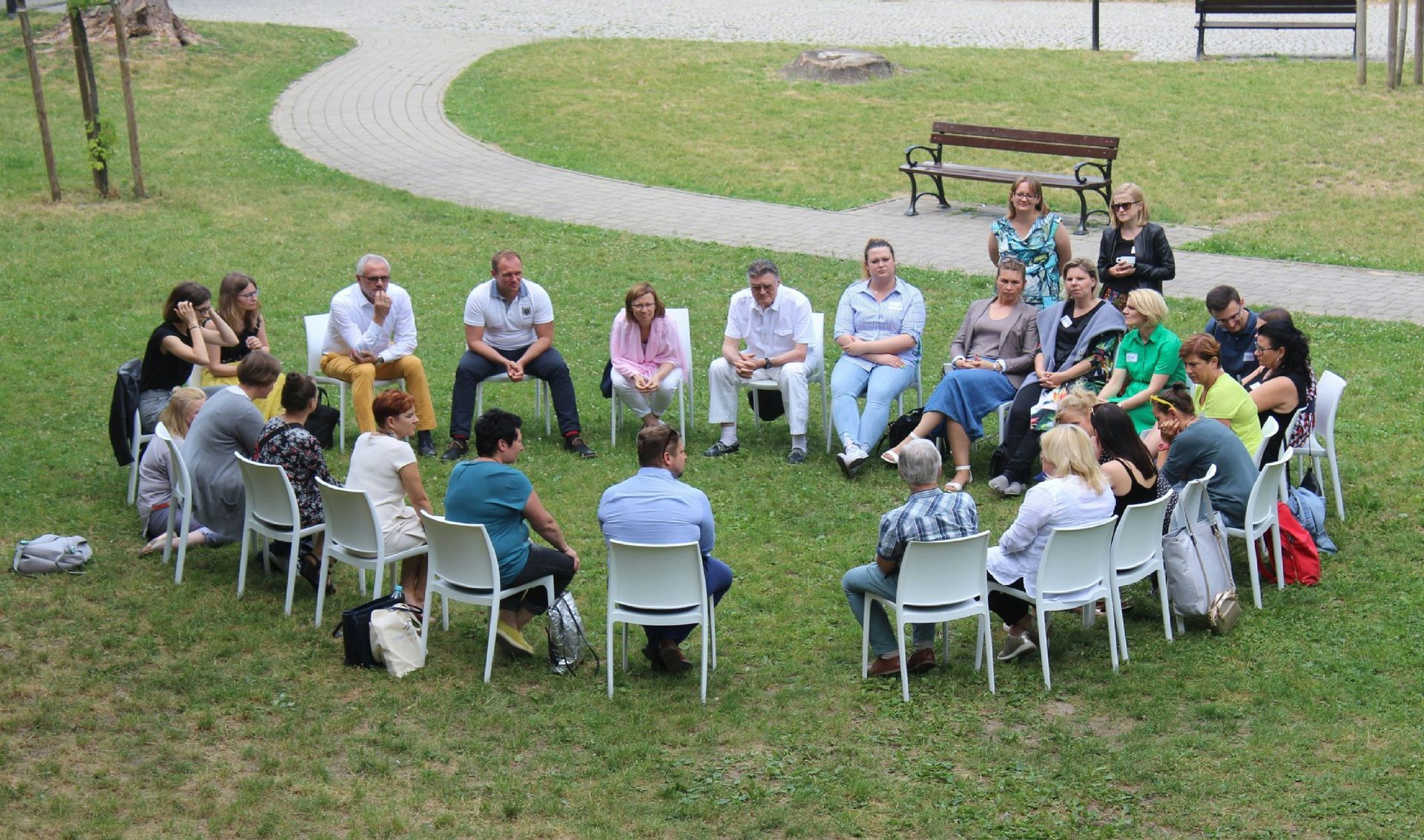 People seated in a circle on grass, engaged in a meeting, with a bench and trees visible in the background.