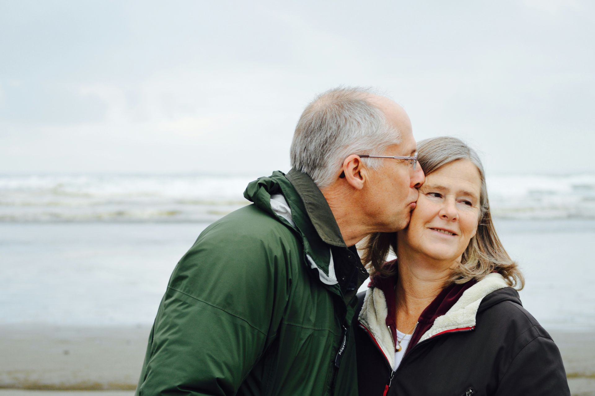 Older couple kissing on beach, man in green jacket, woman in dark jacket, ocean in background.
