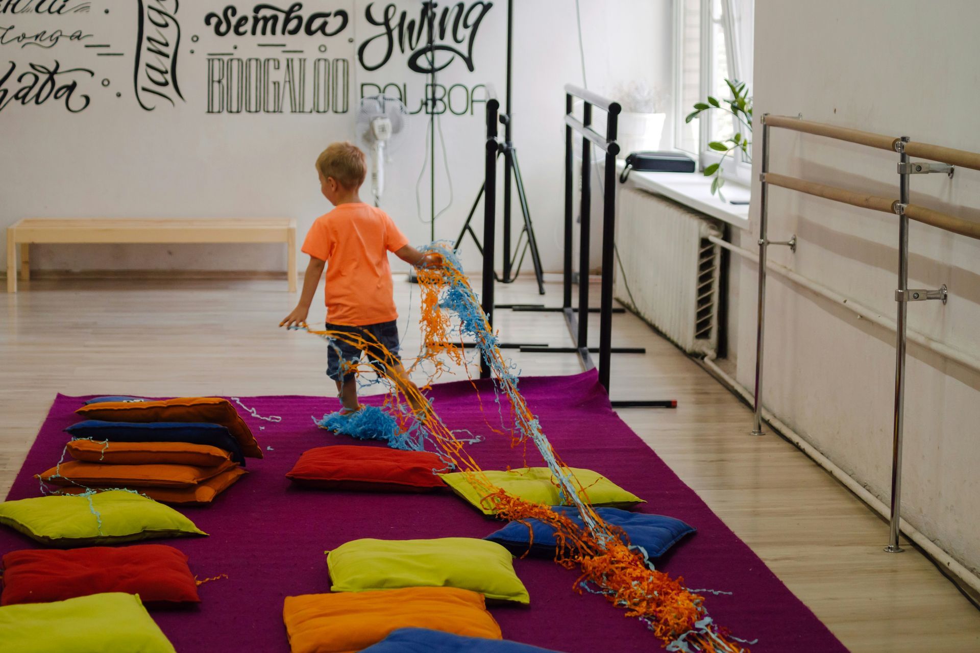A young boy in orange shirt plays on a purple mat with colorful pillows and strings in a dance studio.