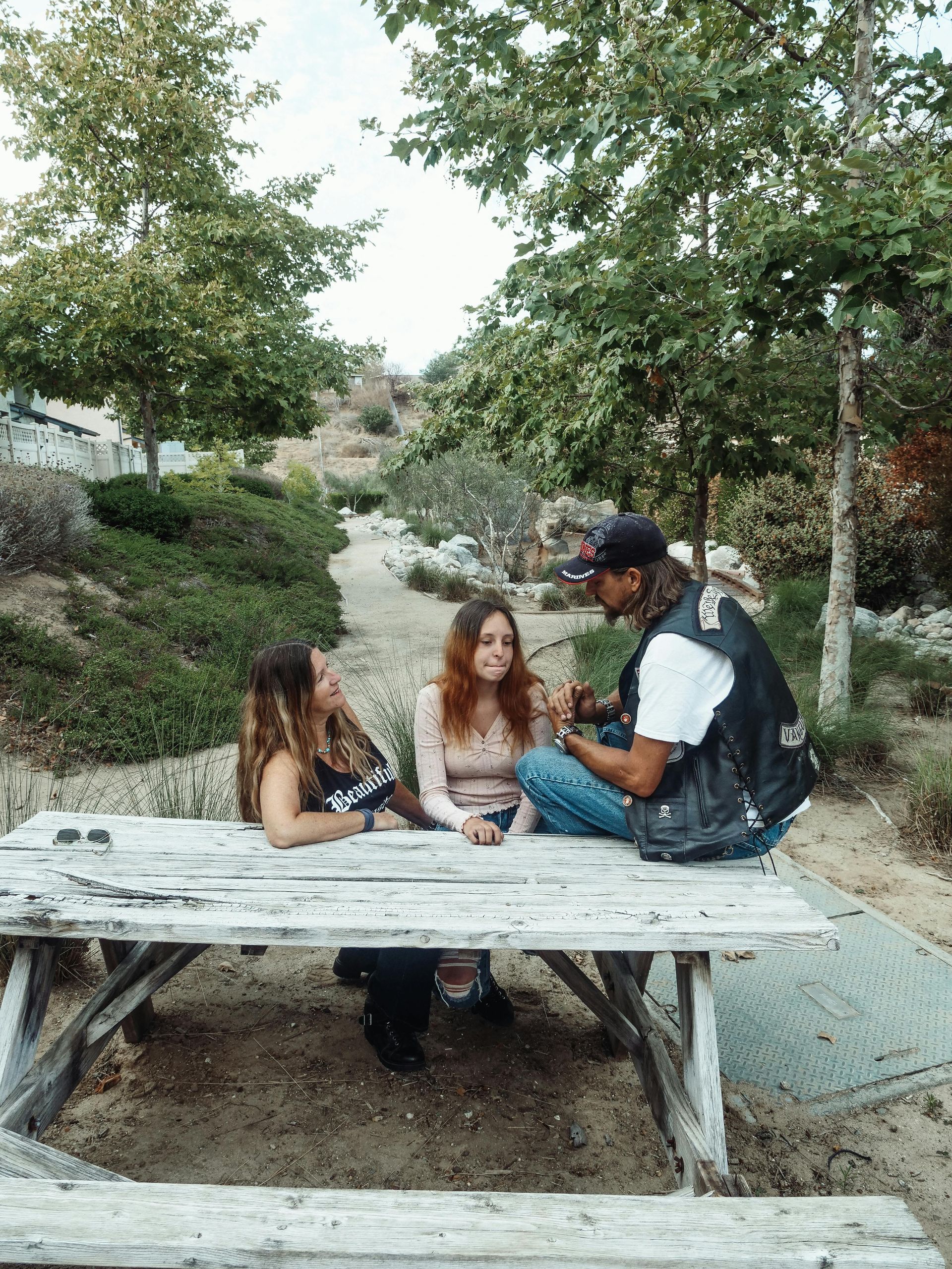 Three people at a weathered picnic table. One adjusts the hair of a woman seated across from him. Park setting.