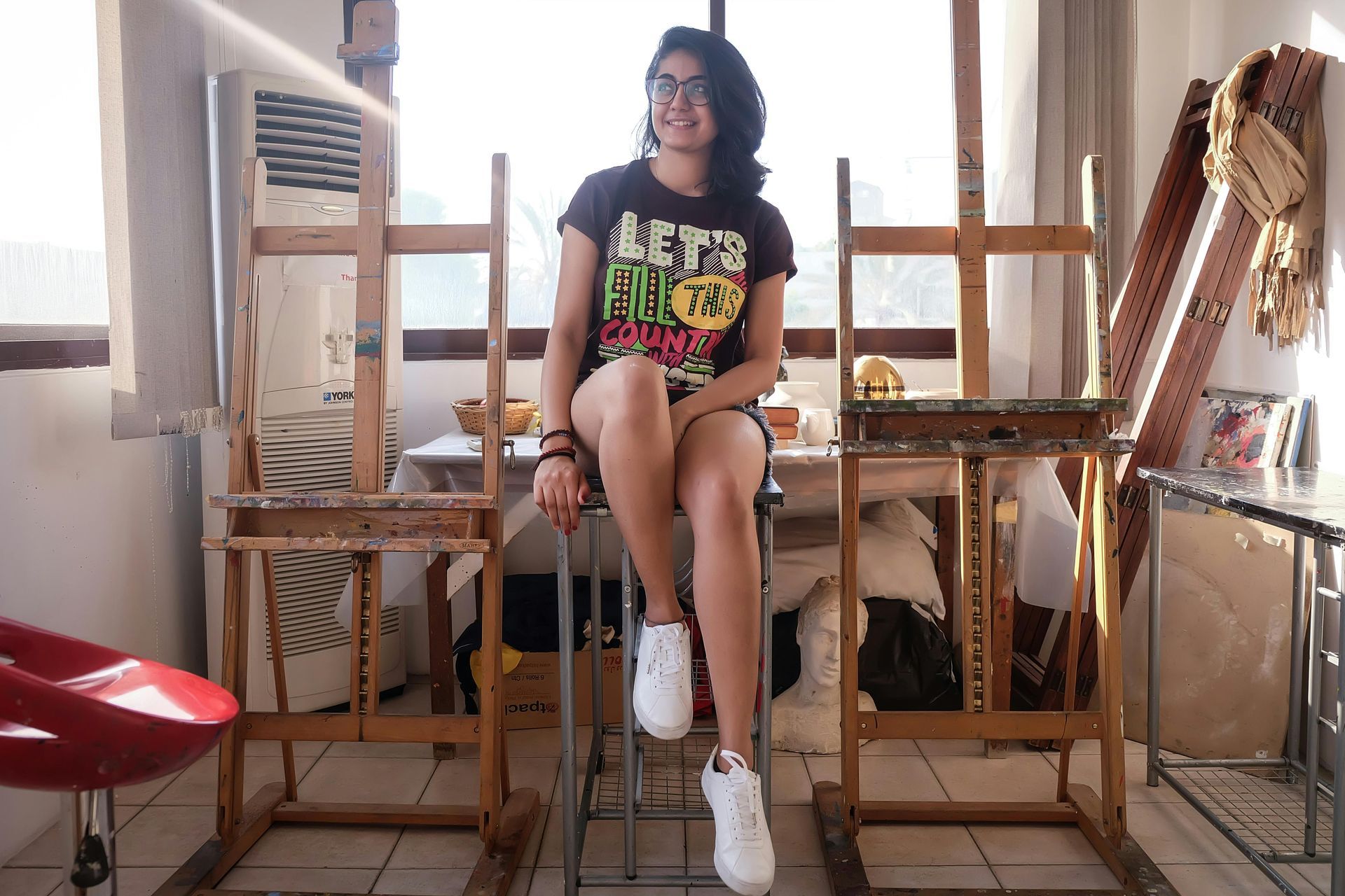 Woman in art studio, sitting between easels, wearing t-shirt, shorts, and white sneakers, smiling.