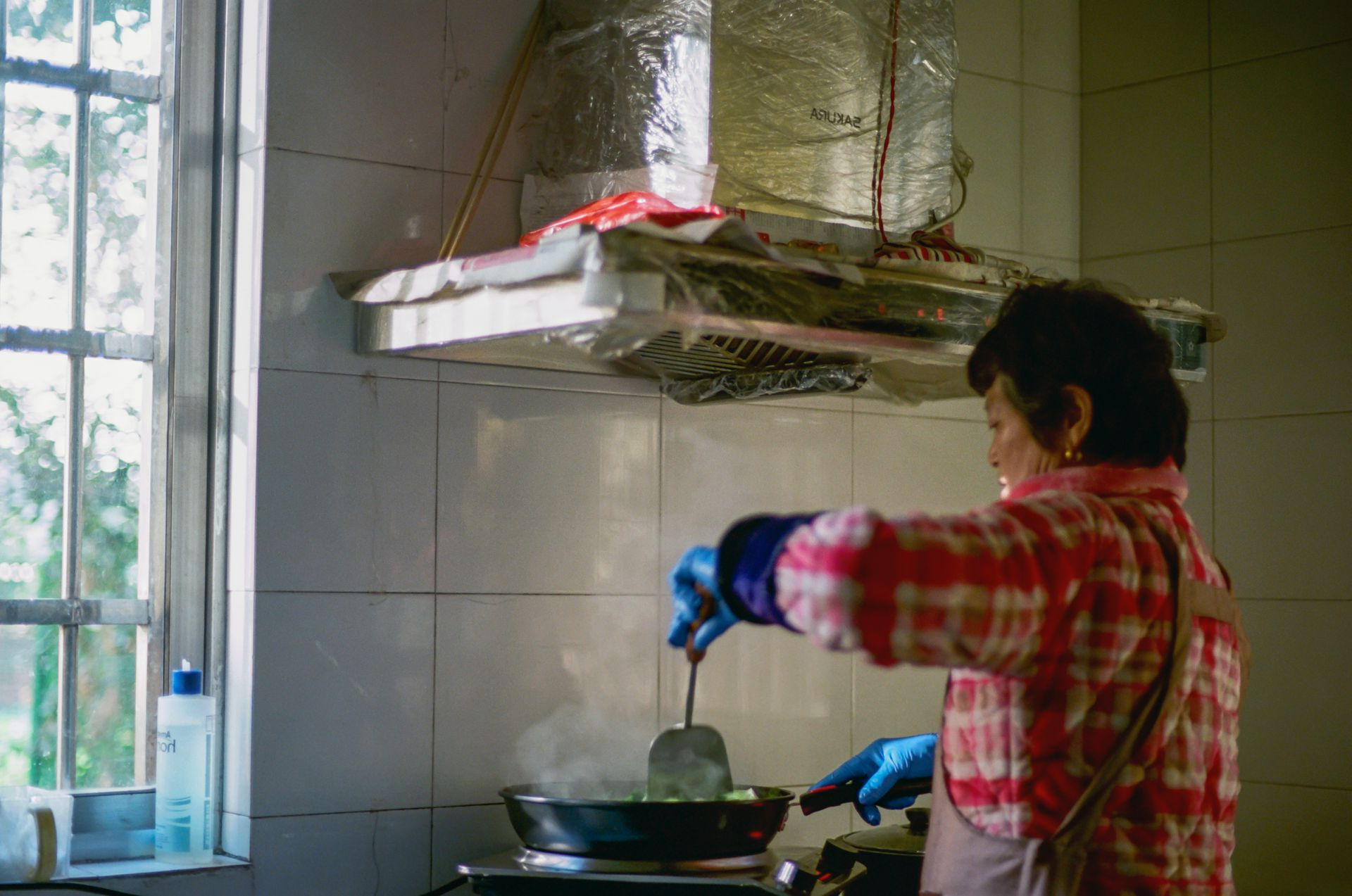 Woman in red plaid shirt cooks in a kitchen, steam rising from the pan.