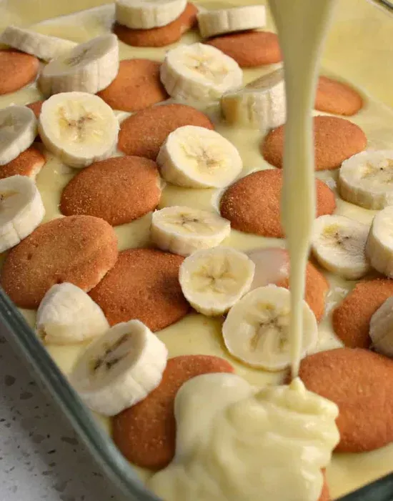 banana pudding is being poured over bananas and cookies in a pan .