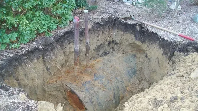 Large pit in the ground exposing layers of earth, pipes, and a shovel.