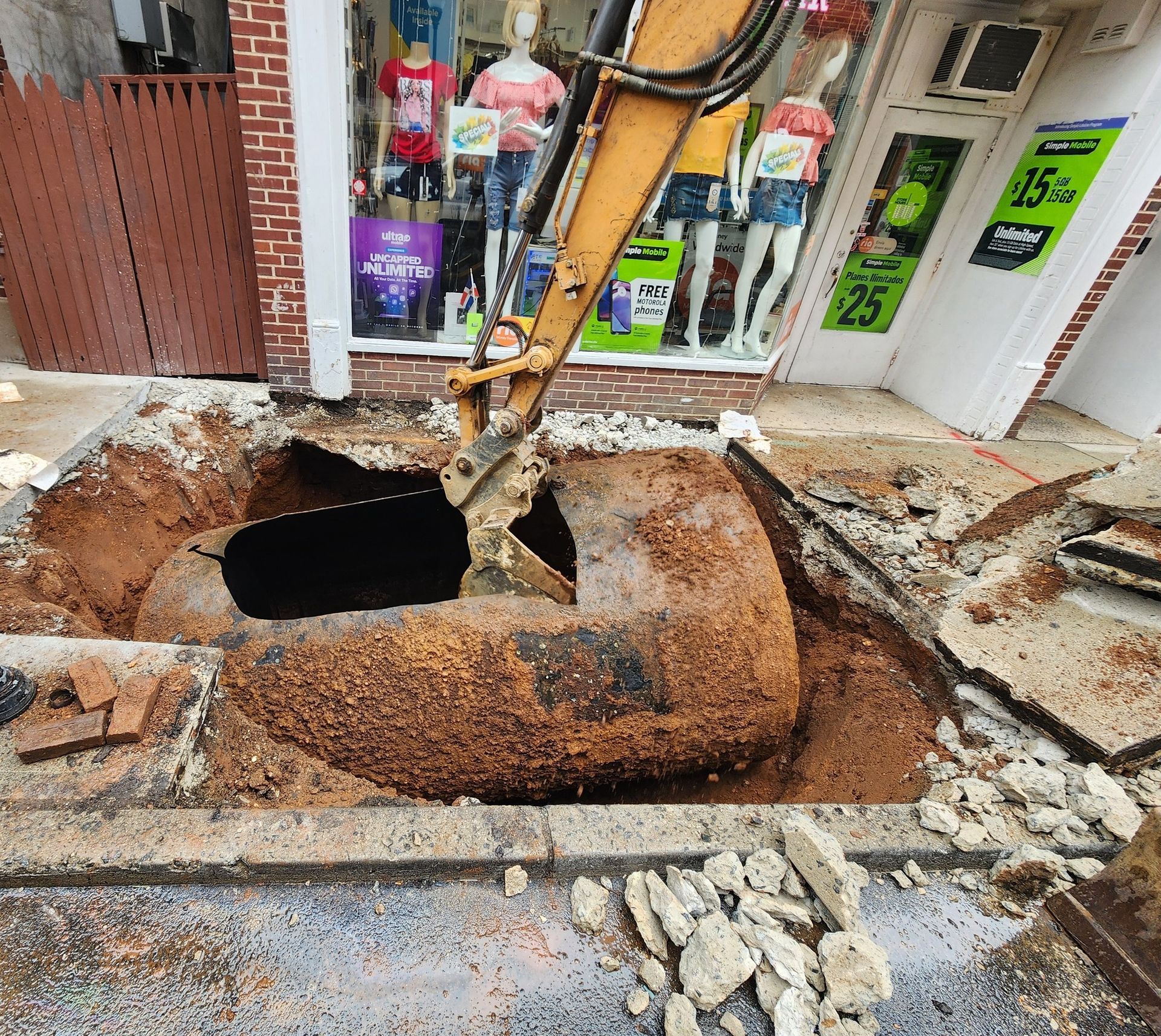 An excavator digs into a sidewalk in front of a store, exposing a large rusted pipe.