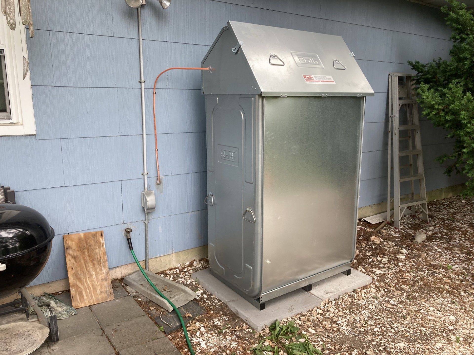 Metal box on a concrete base next to a blue house. A water hose is attached to the house.