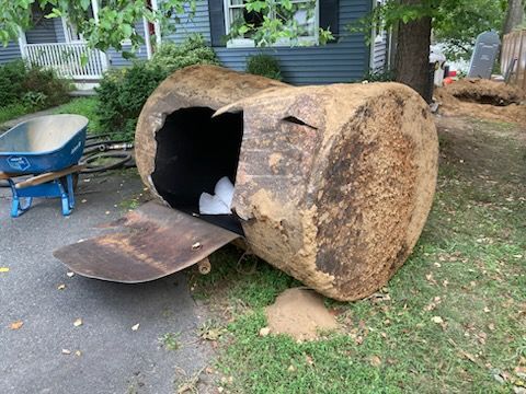 A large, rusty oil tank on the ground with a hole cut in it, in front of a house.