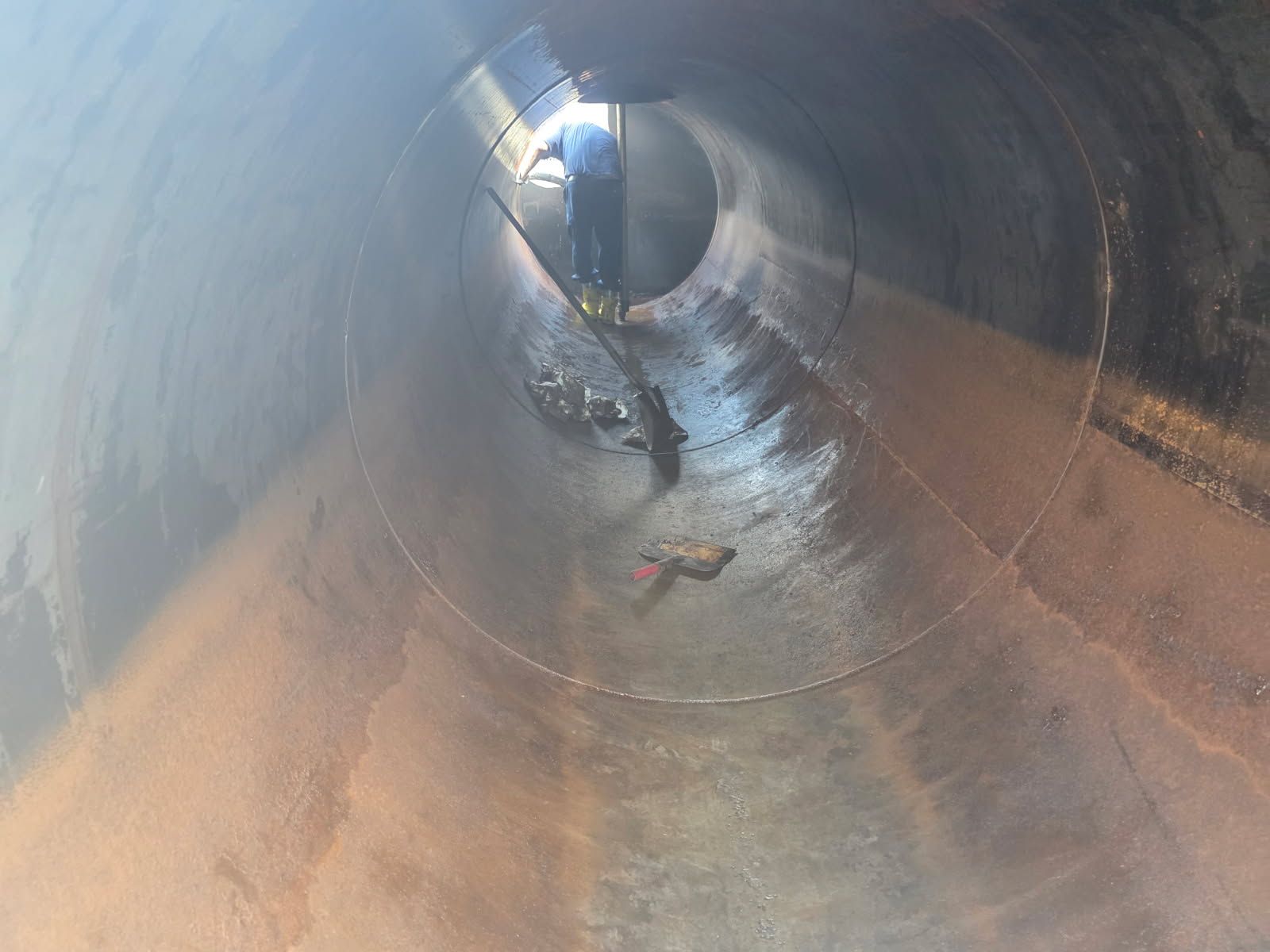 Inside a large metal pipe, a person works, tools visible, with water pooling on the floor.