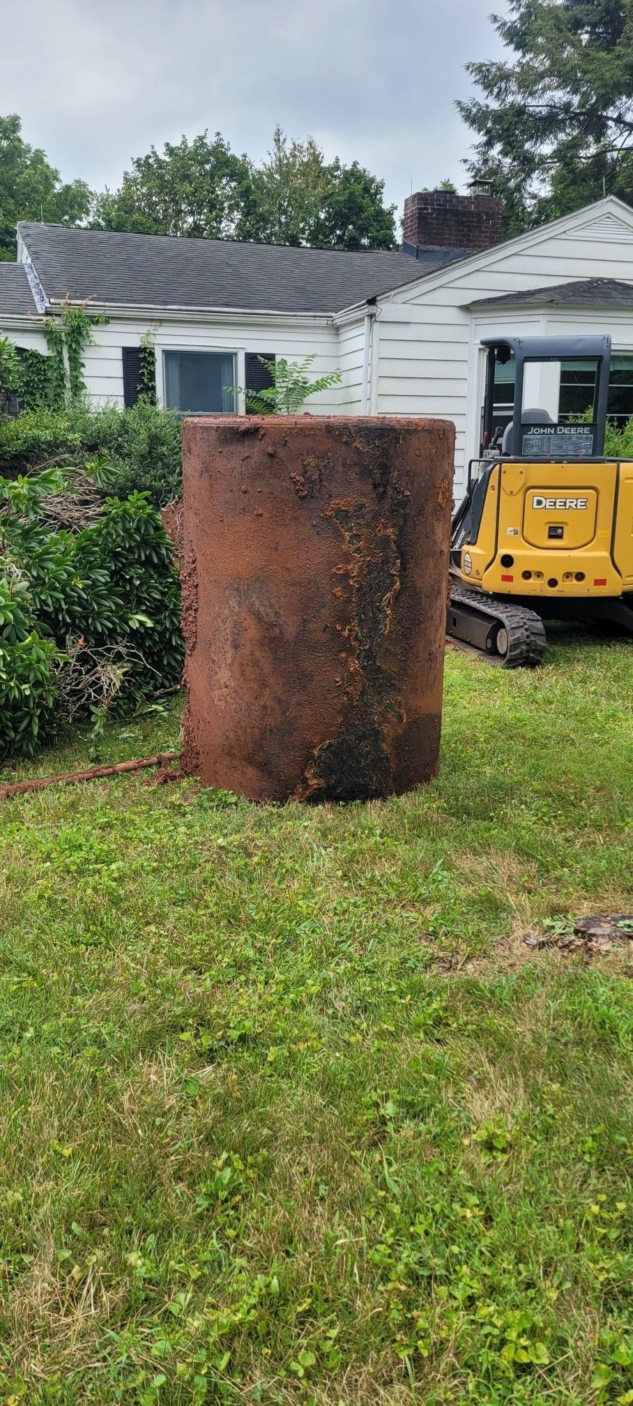 Rusty tank in a grassy yard next to a house and a yellow construction vehicle.