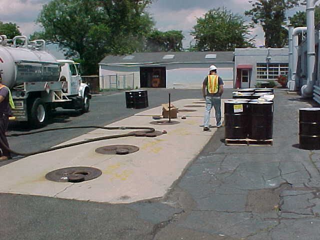 Truck pumping liquid into underground tanks; workers in safety vests, barrels on pallets.