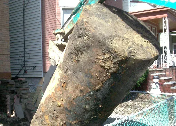 Excavator bucket filled with soil and debris, near a building with a fence.