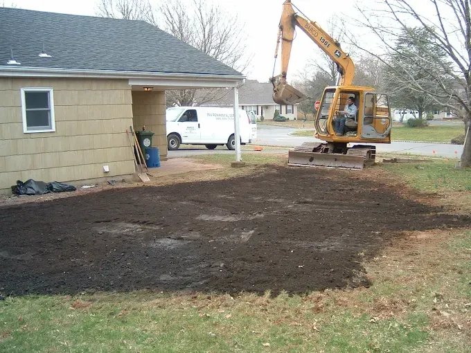 Excavator digging in yard next to house; dark soil patch, white van in background.
