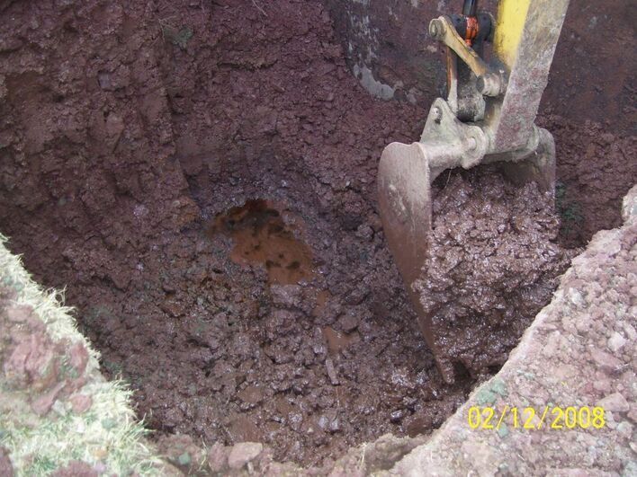 Excavator digging into dark red soil, in an open pit.