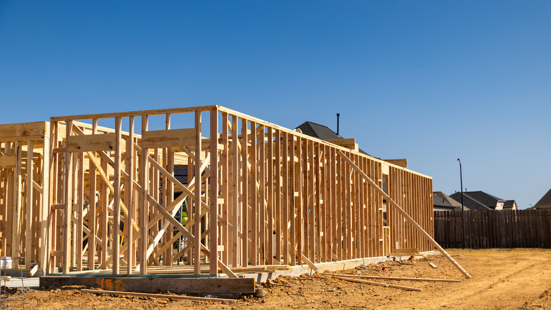 Wood-framed house under construction on a dirt lot beneath a clear blue sky