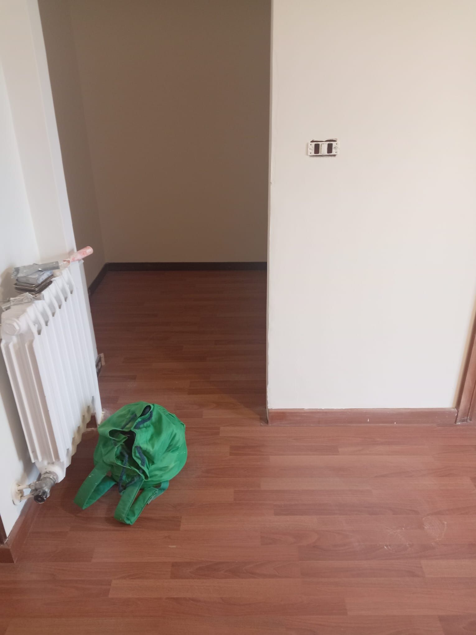 Una stanza vuota con un sacco verde sul pavimento e un termosifone.