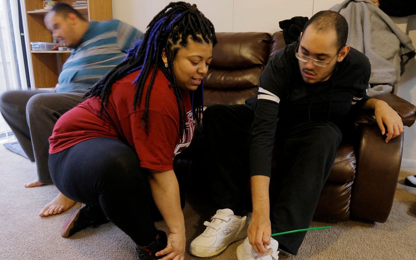 A direct support worker, center, puts on Keith Conley's shoes, while his brother Kristopher moves