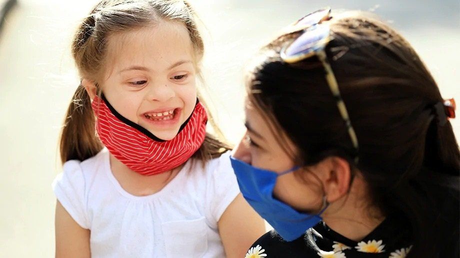 Little girl with Down syndrome smiles at woman in mask.