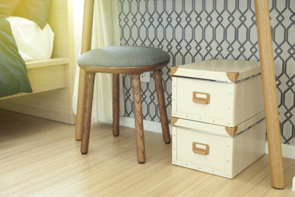 A wooden stool is sitting next to a stack of white boxes in a bedroom.