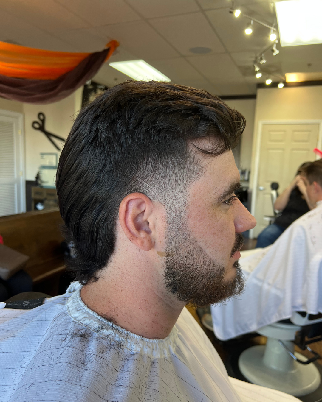 Man with a mullet hairstyle and faded sides, trimmed beard, in a salon.