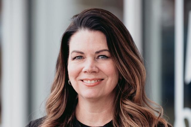 Woman with long, wavy brown hair smiles at the camera, wearing a dark top, outdoors near a building.