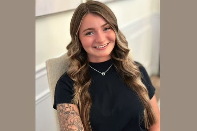 Woman with long wavy hair, smiling, wearing a black shirt and silver necklace. Sitting indoors.