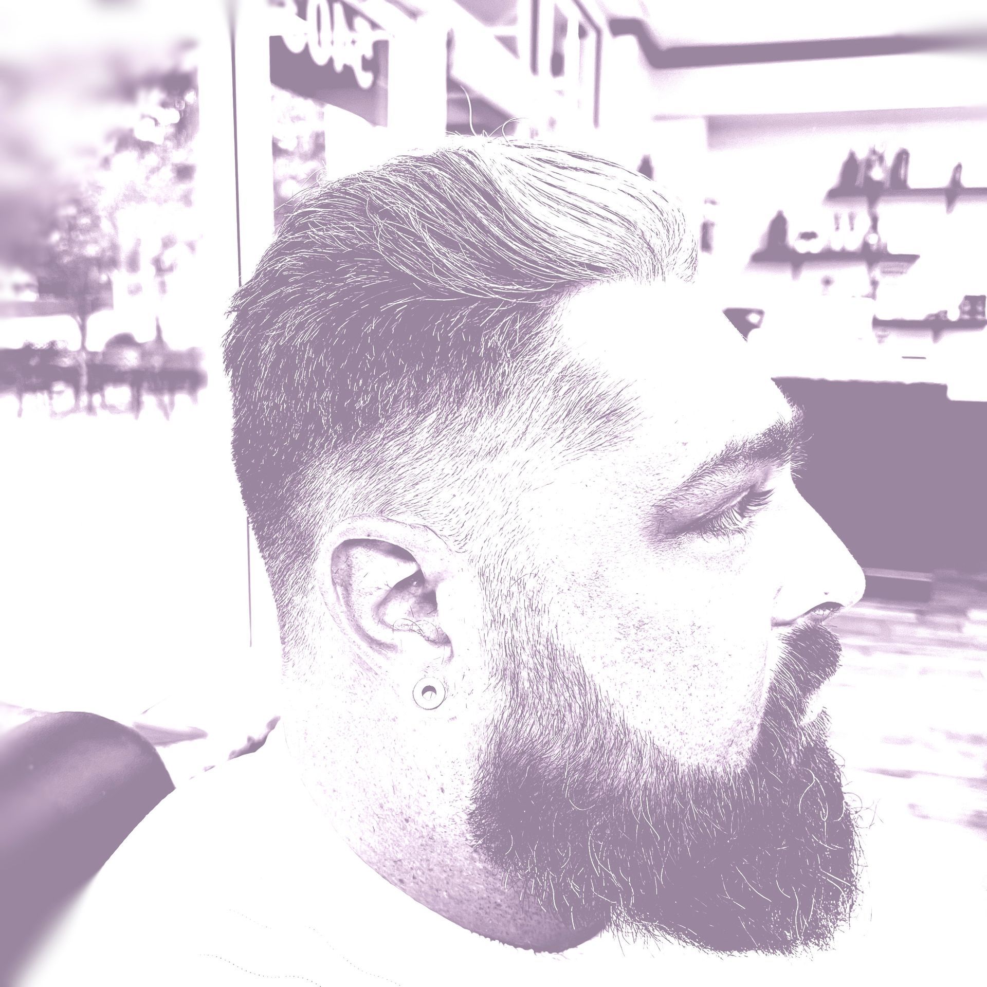 Man with beard, short hair, and ear piercing, looking right. Purple hue, indoor setting.