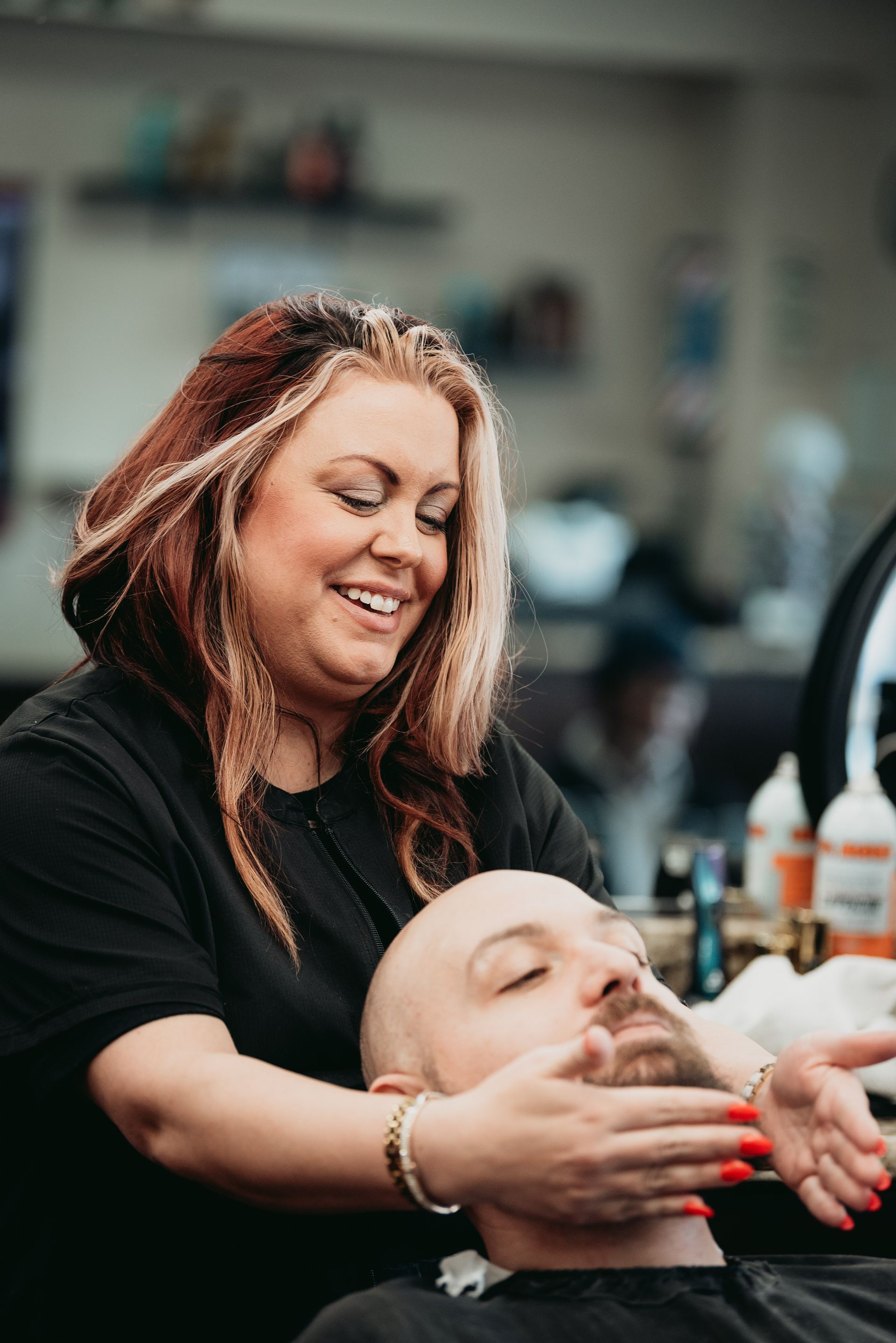 Woman smiling, touching a man's face in a barbershop. He has a beard; she has highlighted hair.