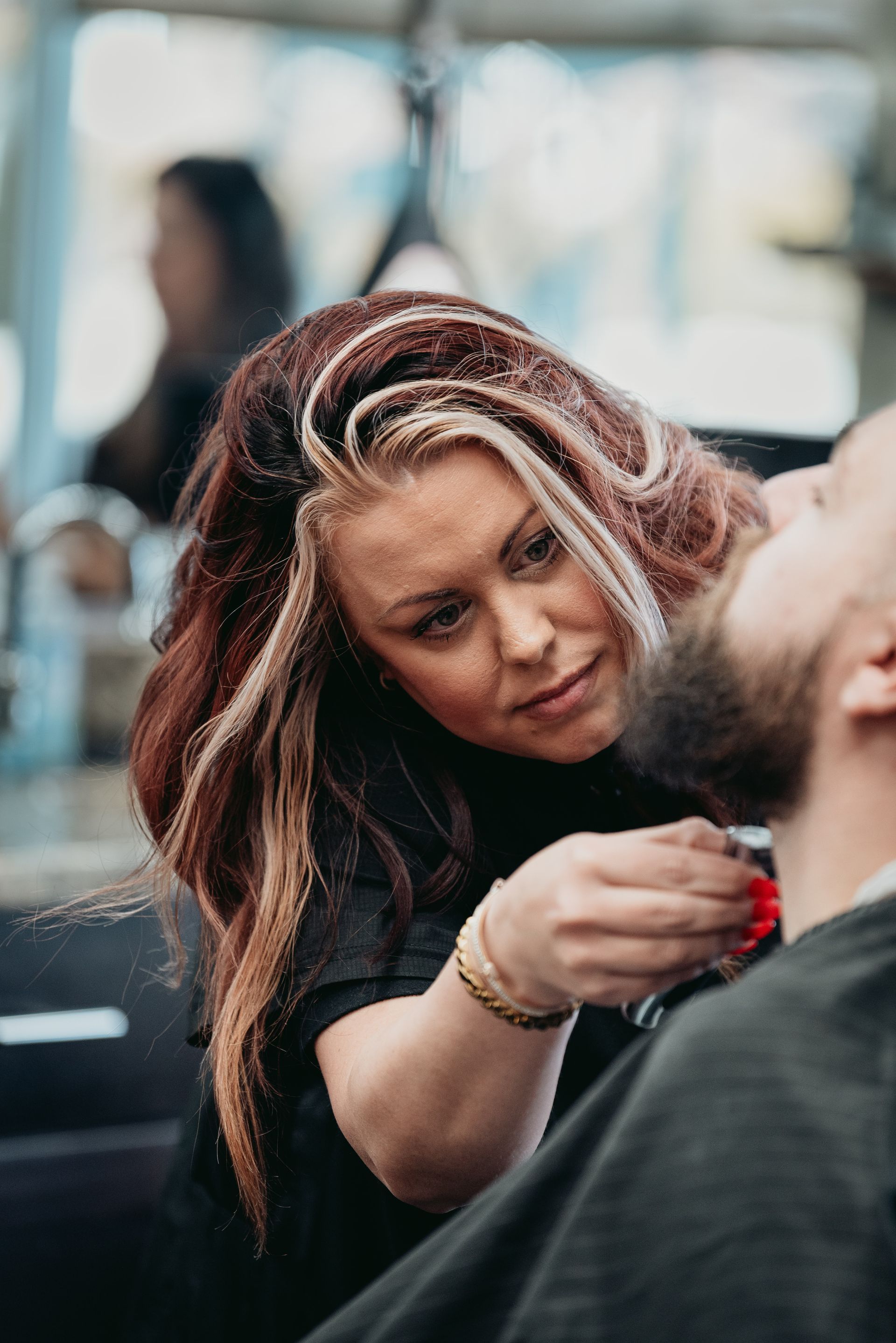 Woman trimming a man's beard with a razor in a barbershop. The woman has red and blonde hair.