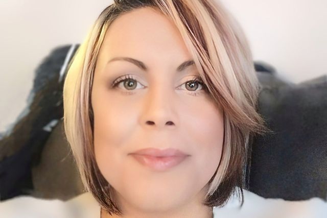 Woman with short, layered hair, light makeup, smiling slightly; against a blurred background.