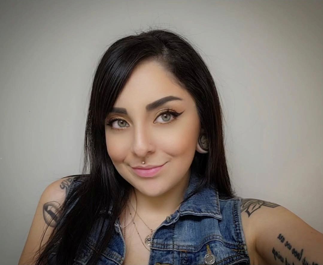 Woman with dark hair, piercings, and tattoos smiles, wearing a denim vest, and looking at the camera.