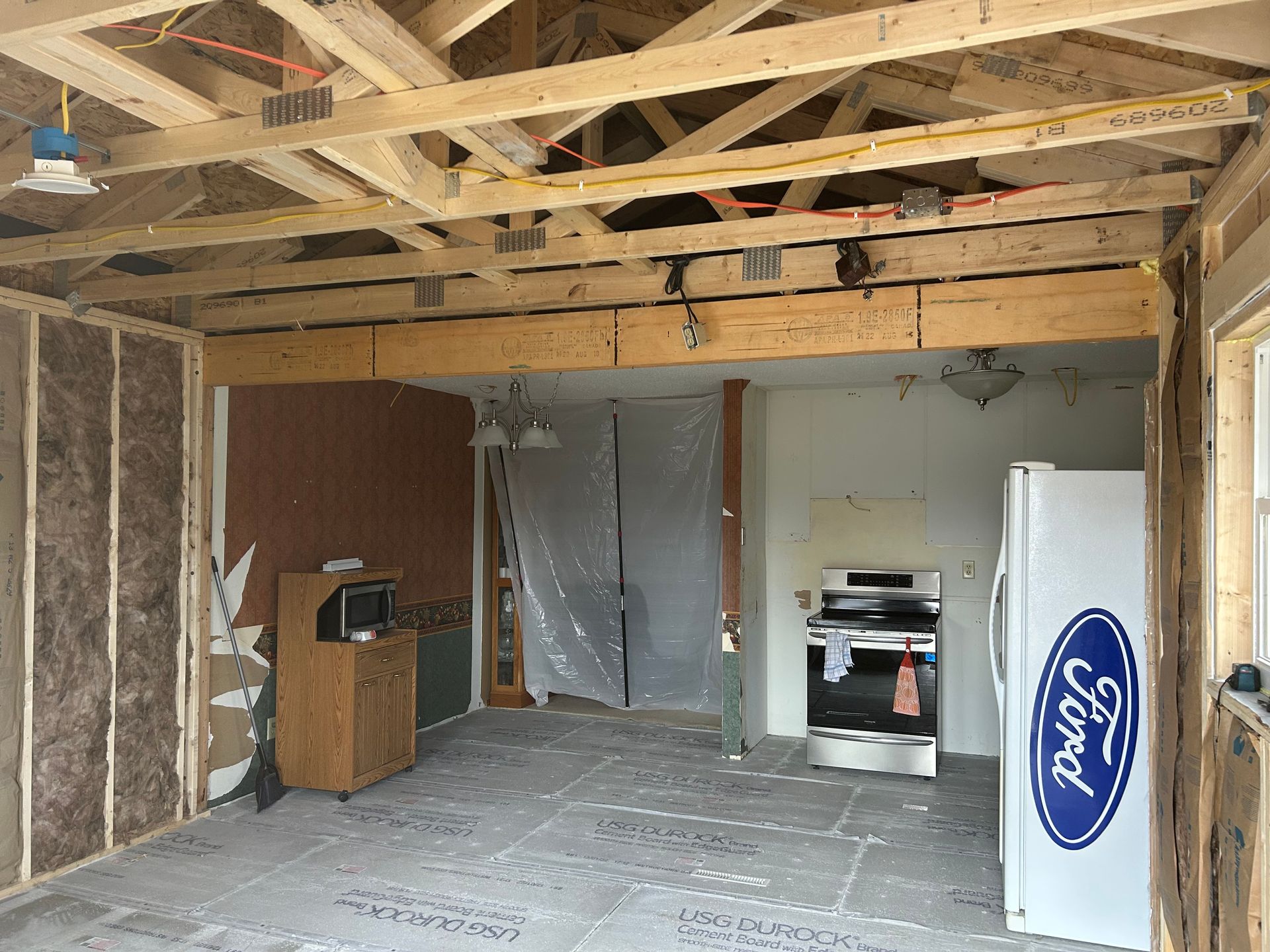 Interior framing of a room under construction with exposed wooden beams, insulation, appliances, and a plastic sheet.