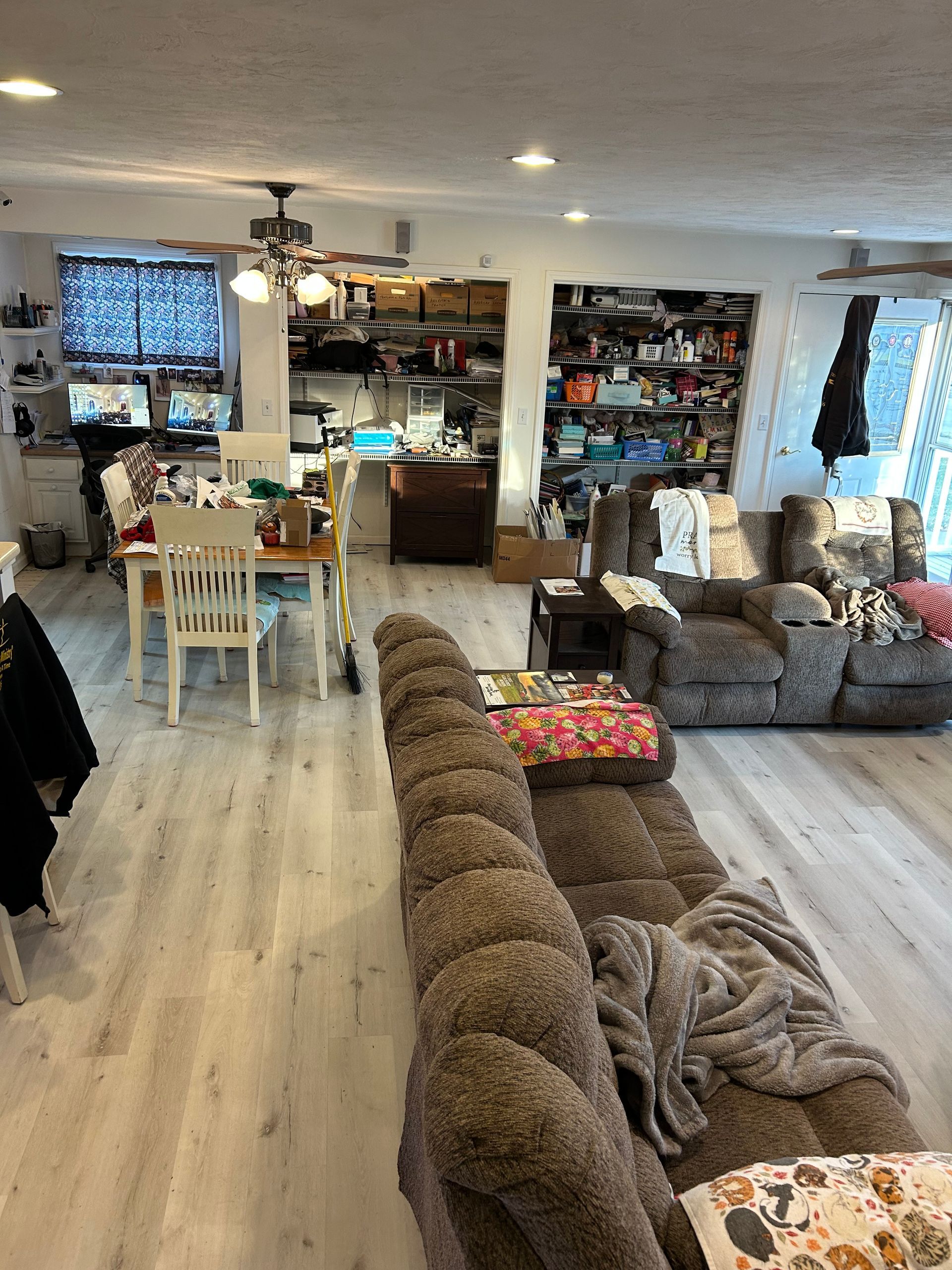 Living room with brown reclining sofa, dining table, and cluttered shelves in the background.