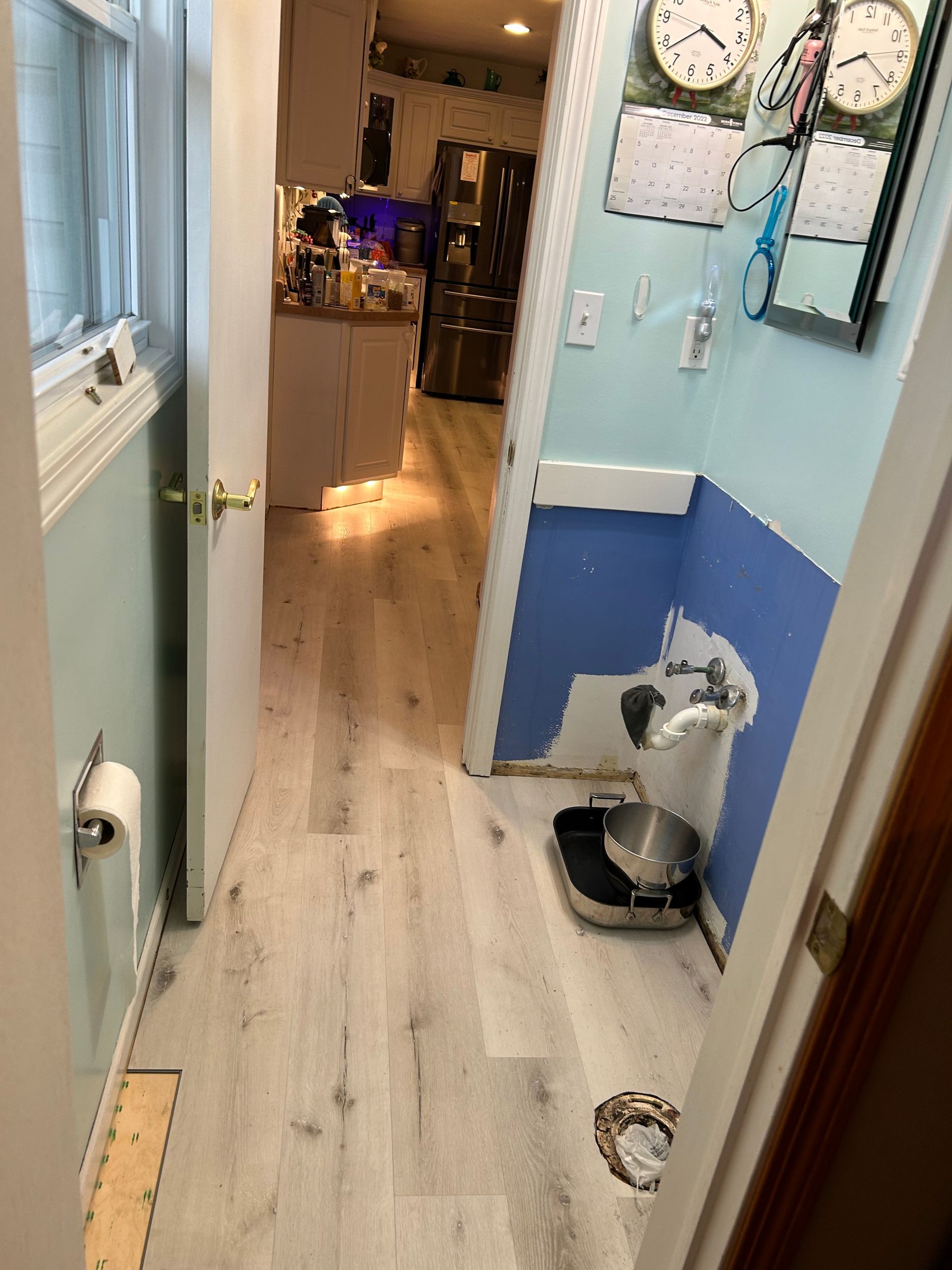 Narrow hallway with new light wood flooring. Bathroom is at the end with blue wall paneling, a scale, and toilet.