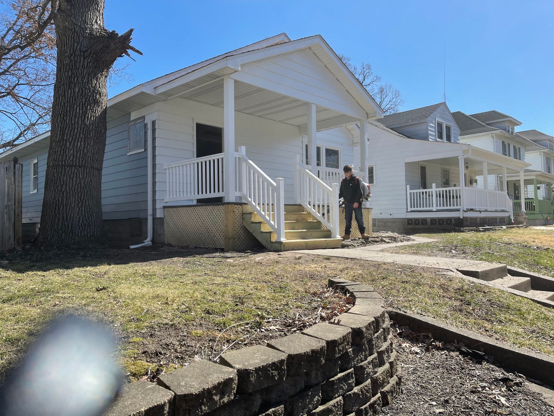 Person standing on the porch of a white house with a covered porch and white railing, on a sunny day.