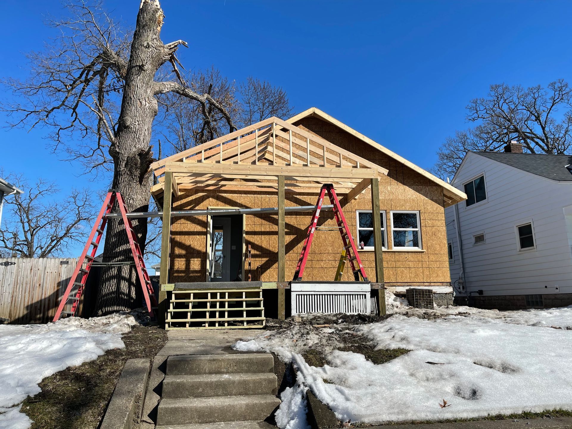 House under construction with porch and roof framing; ladders present, snow on ground.