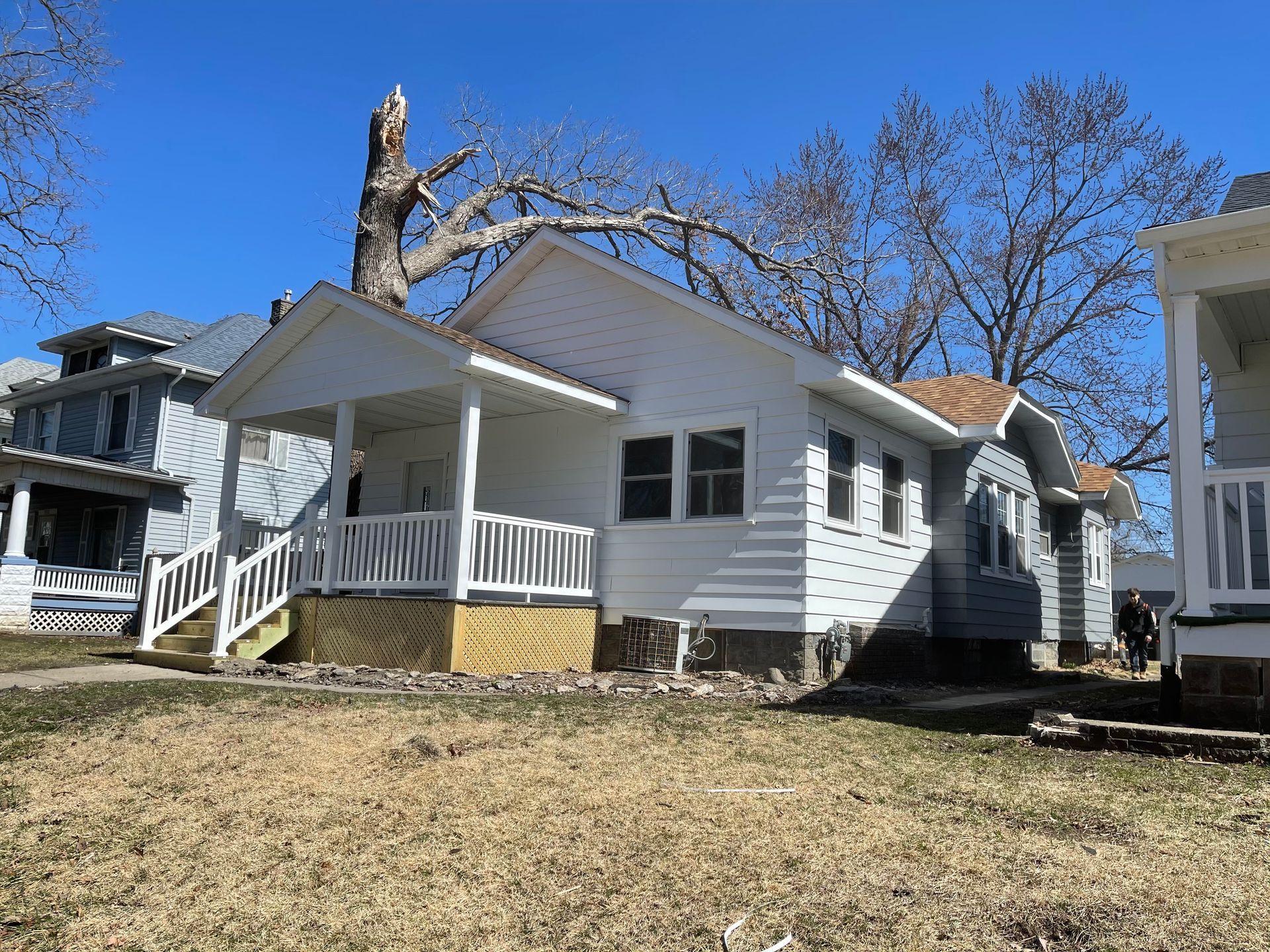 White house with porch, brown roof, and leafless trees against a blue sky.