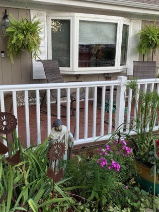 White porch with bay window. Hanging ferns, seated chairs, statue, and flowers.