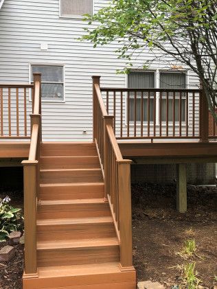 Wooden deck with stairs leading up to it, next to a house with gray siding.
