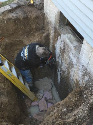 Person using power tool on a concrete foundation wall in an excavated area. Earth, ladder, and a window are visible.