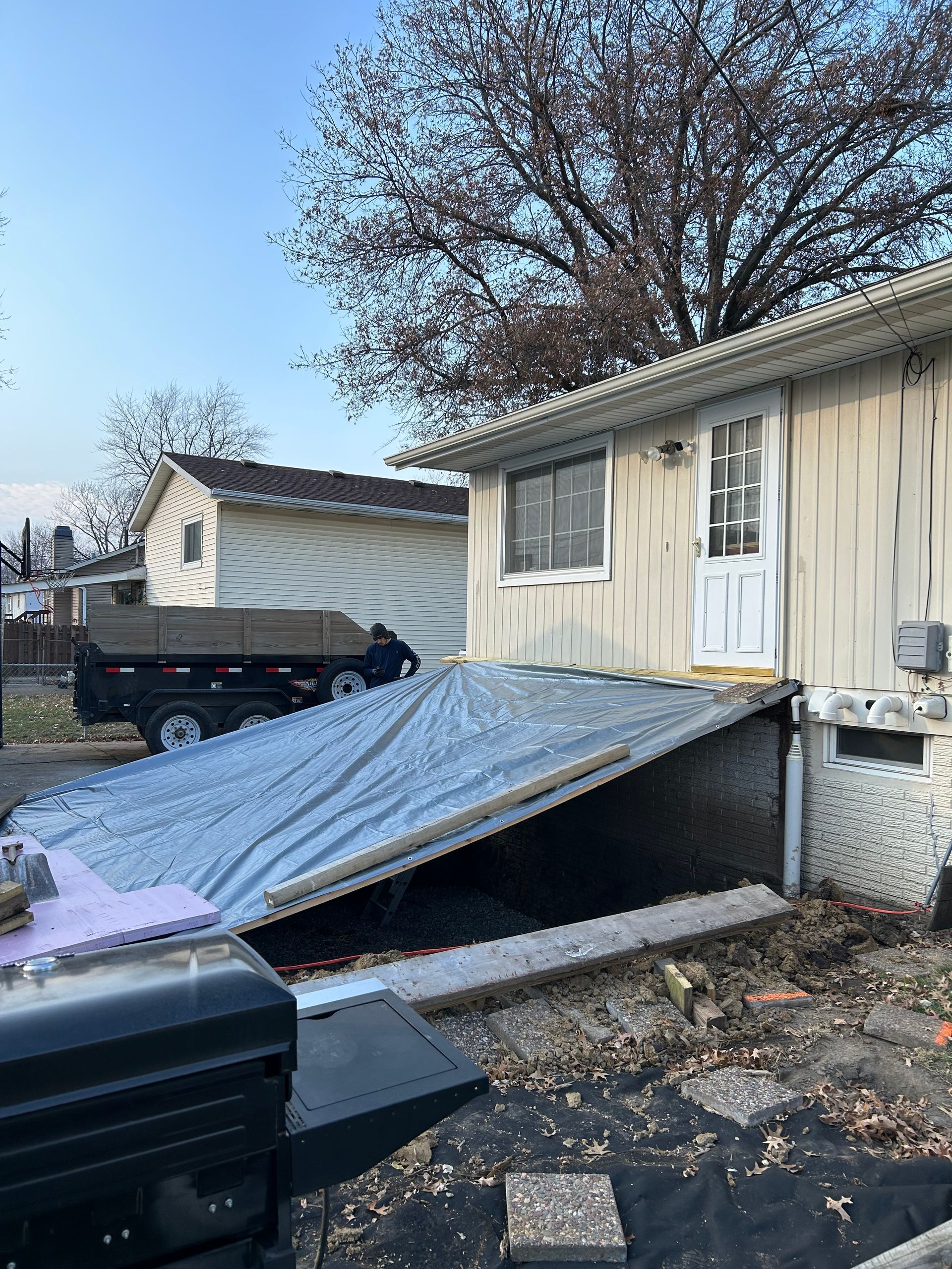 A house with a slanted entryway covered by a tarp, a small window, and a white door. Dump truck is behind.