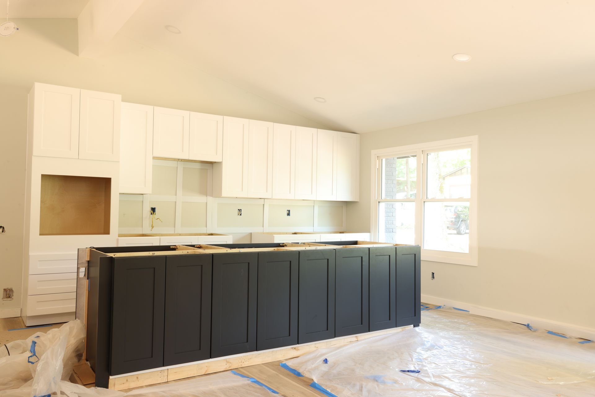 Kitchen under construction with white upper cabinets, dark island cabinets, and a window.