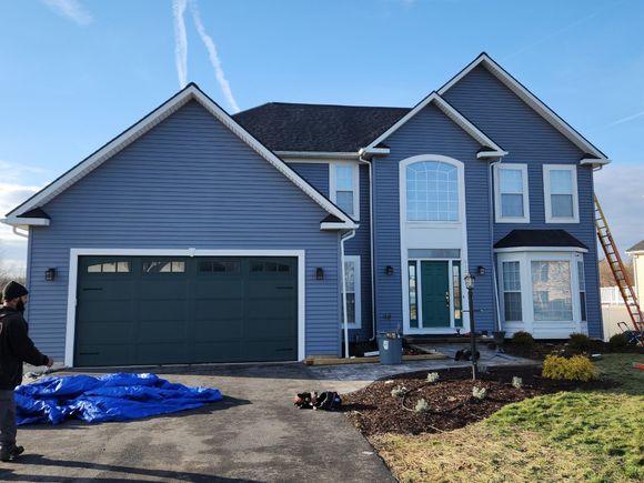 A man is standing in front of a large blue house with a green garage door.