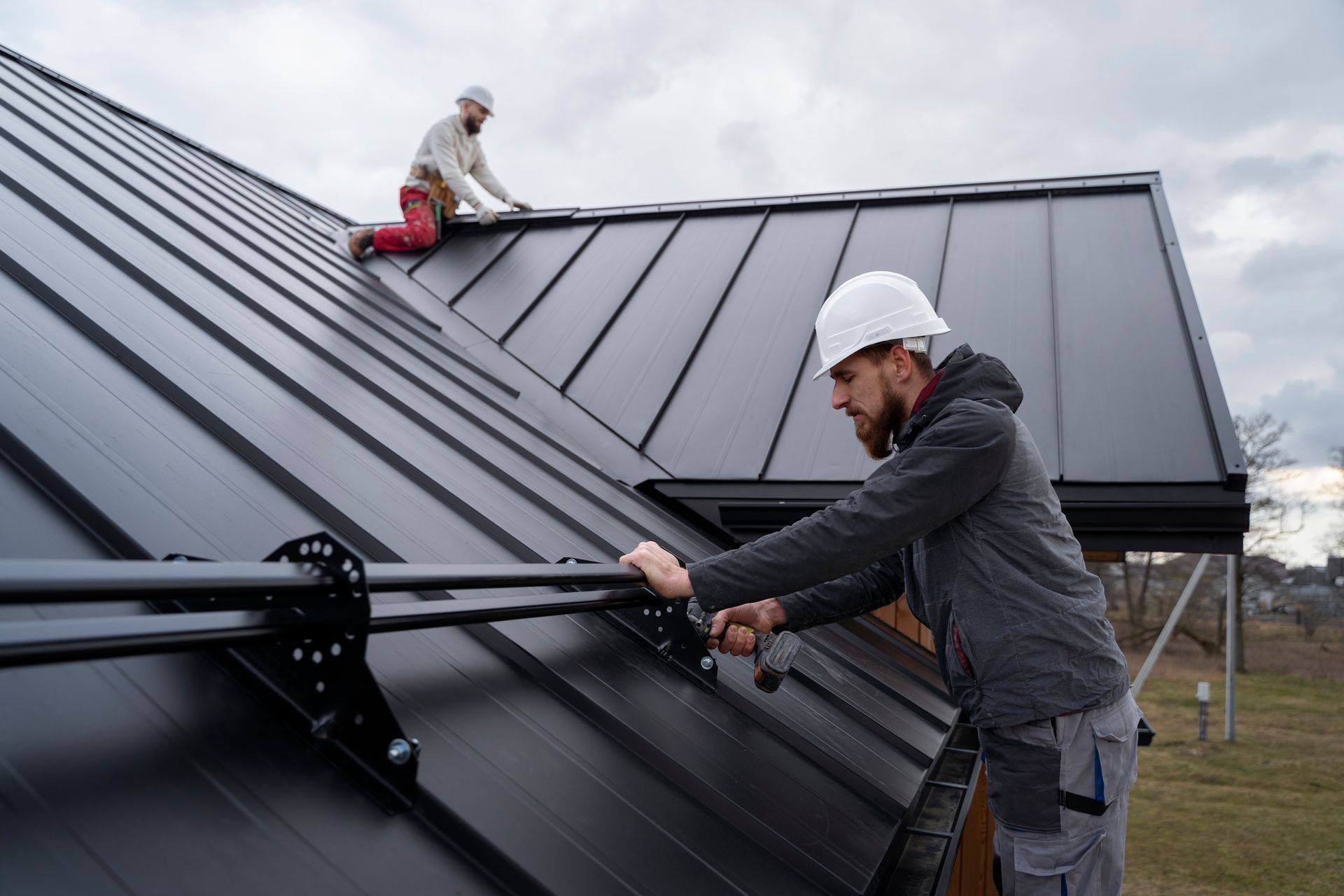 Two roofers installing metal roof panels on a house; one on the roof, one on scaffolding.