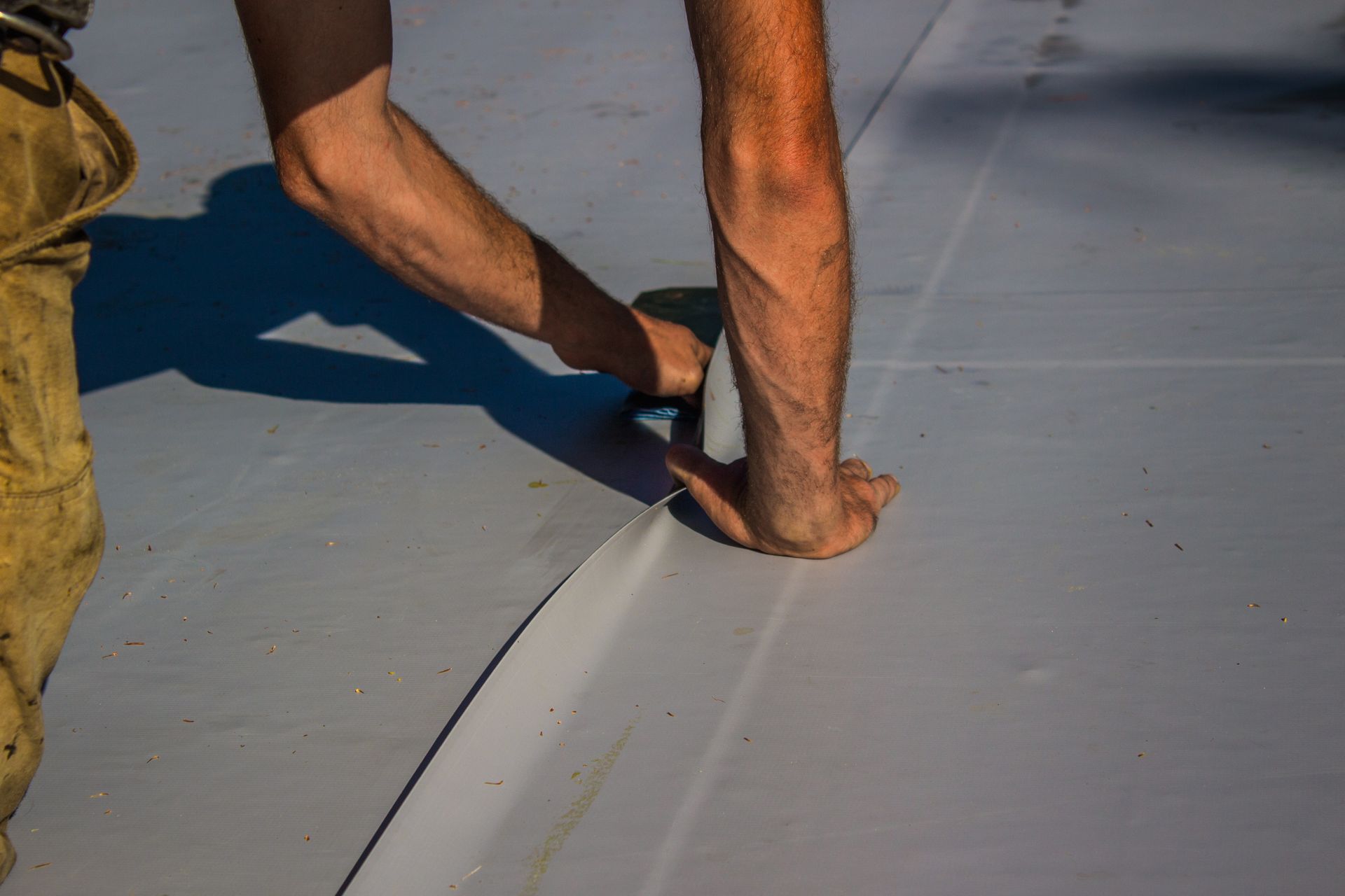 Person using a tool to smooth the edge of a gray roofing material on a sunny day.